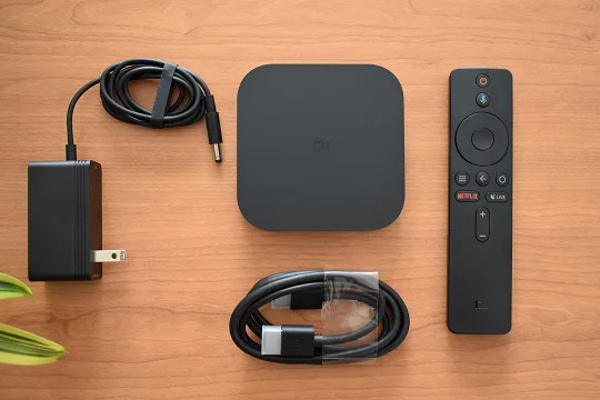 The image shows the Mi Box S 4k packaging content: the box tv, the remote control, the cable HDMI and a charger.
